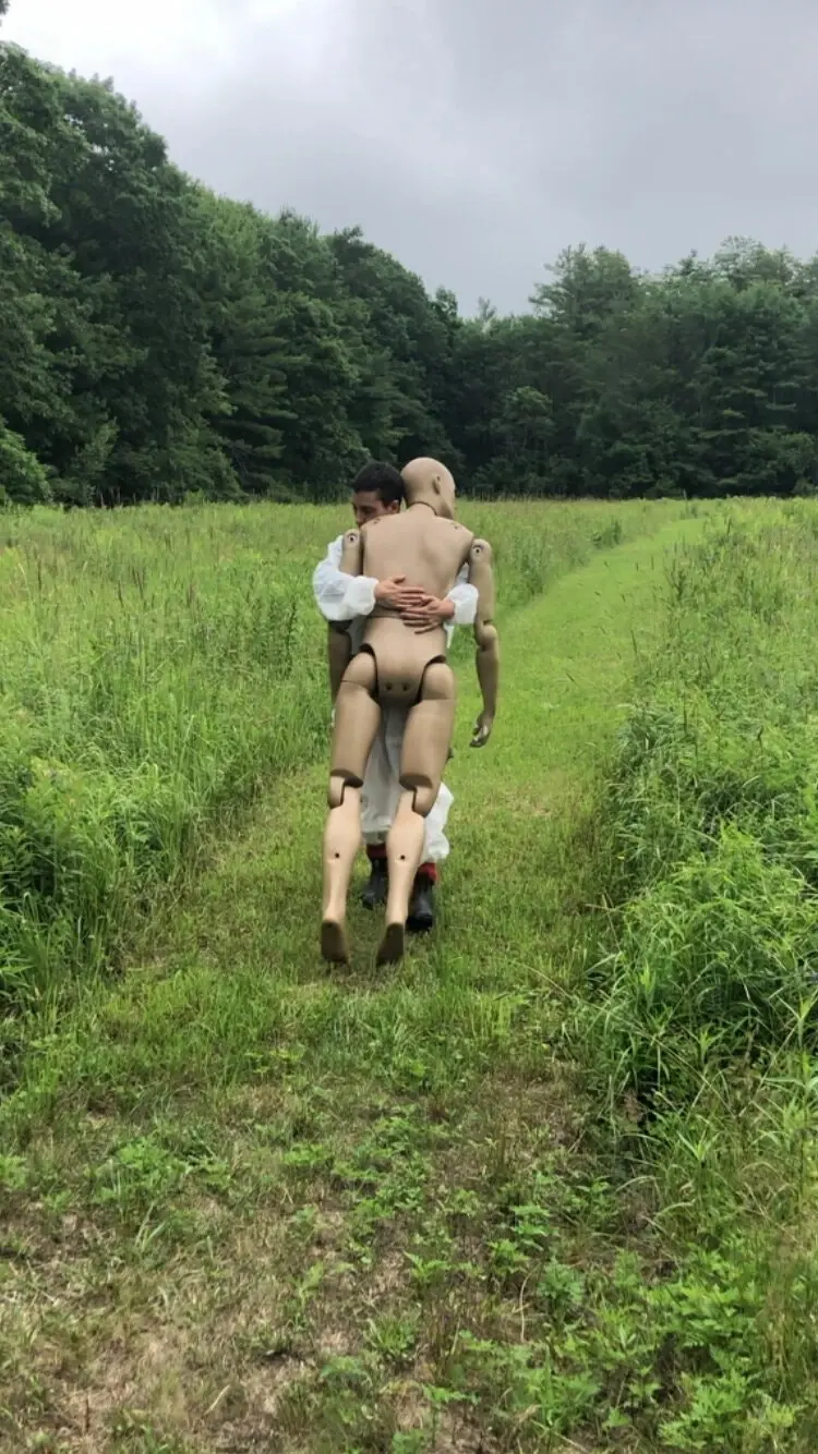 devynn emory from NYT article March 25 2021_6, shows emory walking in a grassy field in a path in a white jumptuit hug-holding a life-sized wooden mannequin.jpeg