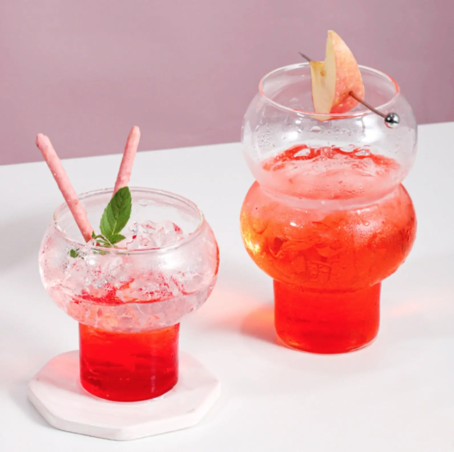 two bubble-glasses holding icey red drinks on a white table, pink wall in the background