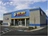 Outside corner view of Napa Auto Parts store on a sunny day. The building is metal and concrete with grey, clue, and yellow coloring and the Napa Logo.