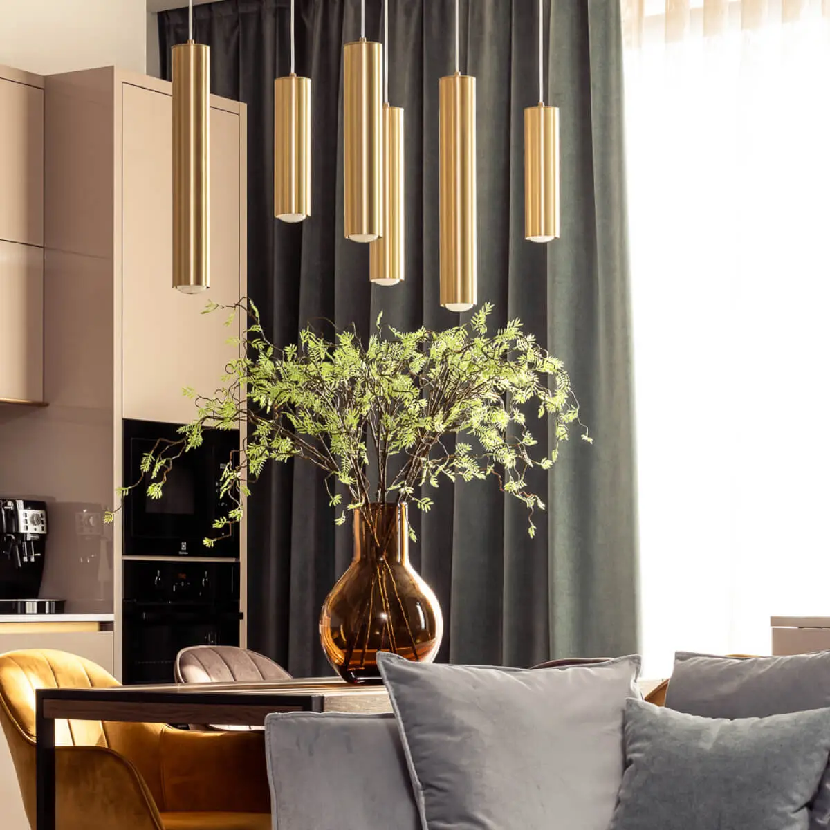 Six cylindrical golden light pendants hang above a dining table. The table has a large orange glass vase full of green foliage. There's velvet green curtains in the background and a light blue sofa in the foreground.