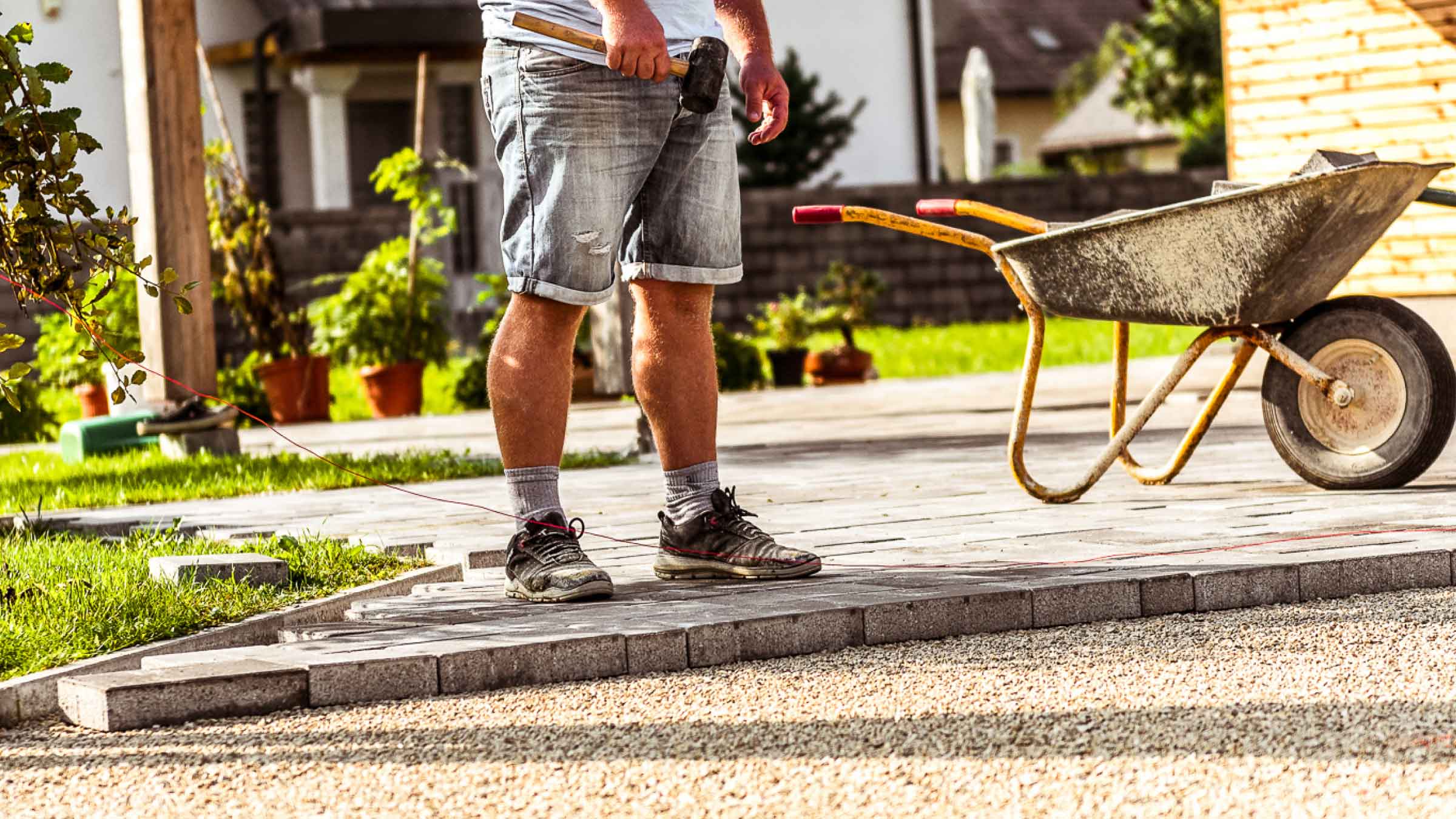 Tradie stands in driveway with rubber mallet in hand. Wheelbarrow by his side and a half finished drive way in front of him.