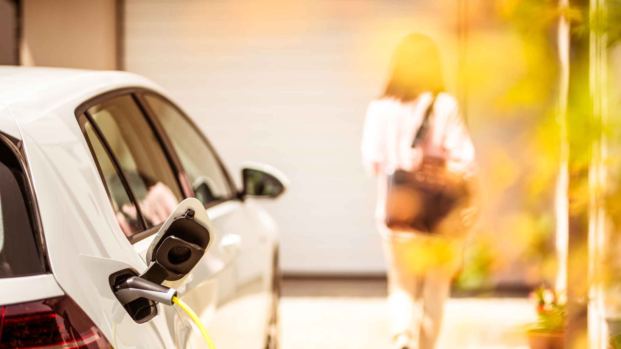 Woman walks toward her house leaving her car plugged in and charging in the driveway behind her.