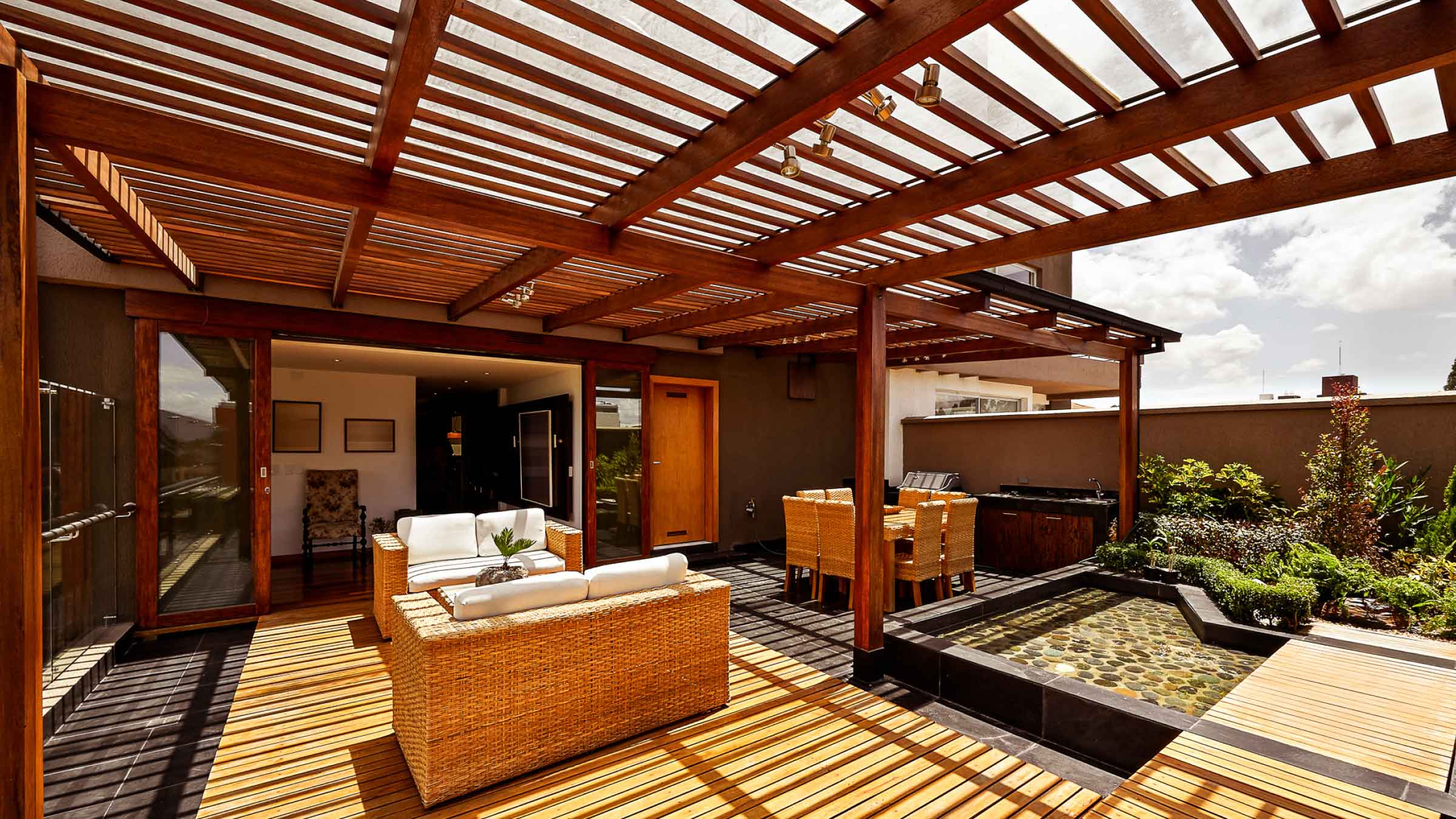 Wooden structure on outdoor entertainment area, roof has shutter blinds that are open, casting shadows on the floor.