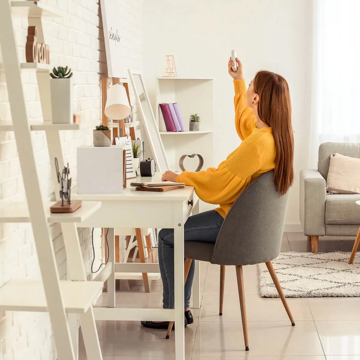 Young woman with long red hair aims her remote control up at the air conditioner in her modern studio apartment.