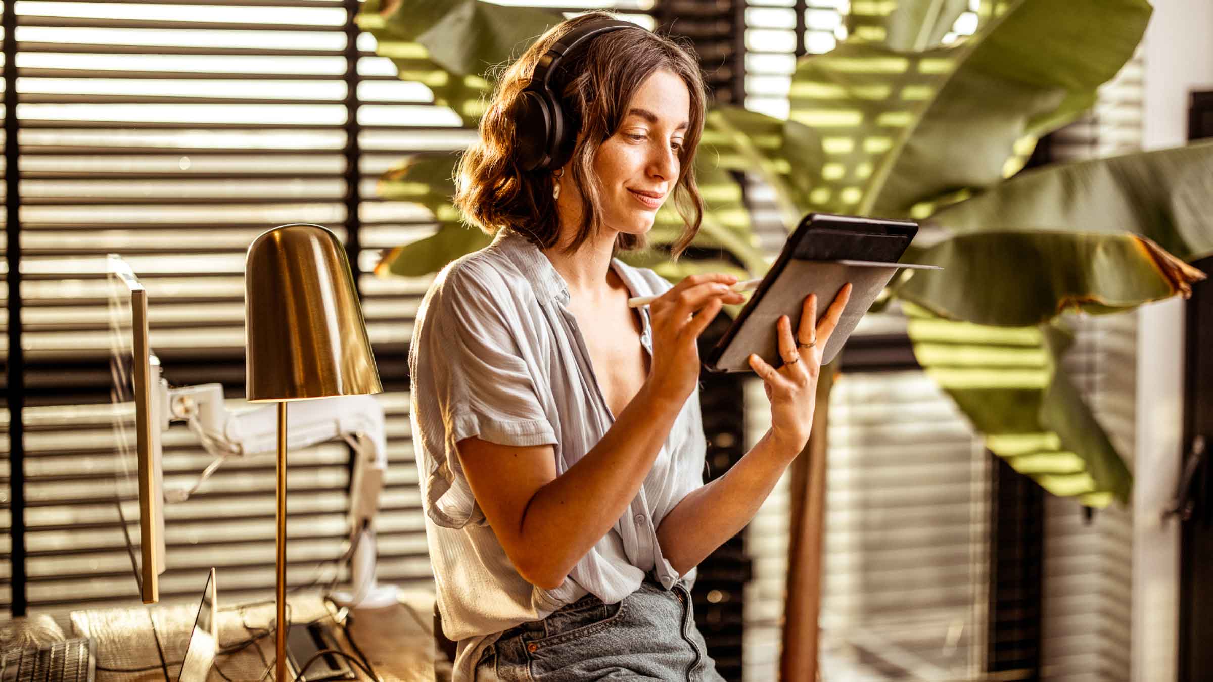 Young lady with headphones on and smiling at her iPad, leans on a table. Window blinds fill the background, and cast shadows onto the leafy green plants in her living room.