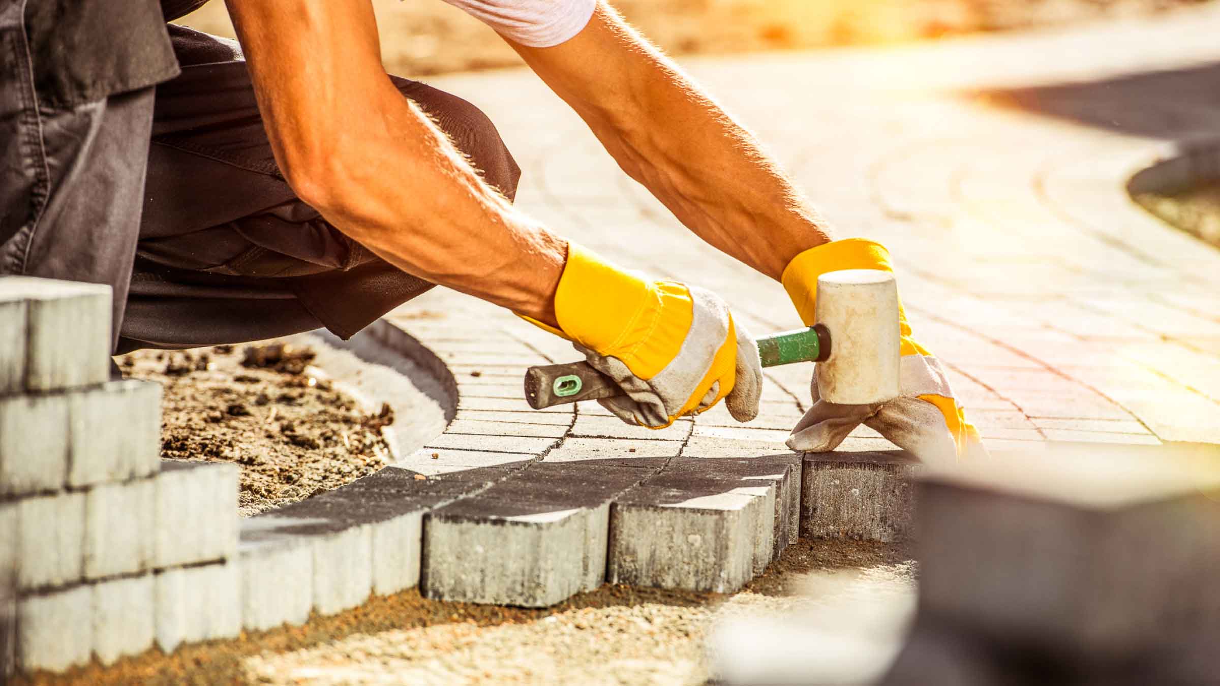 A tradie wearing yellow gloves kneels beside a half finished path. He is laying brick pavers down using a rubber mallet to secure them in place.