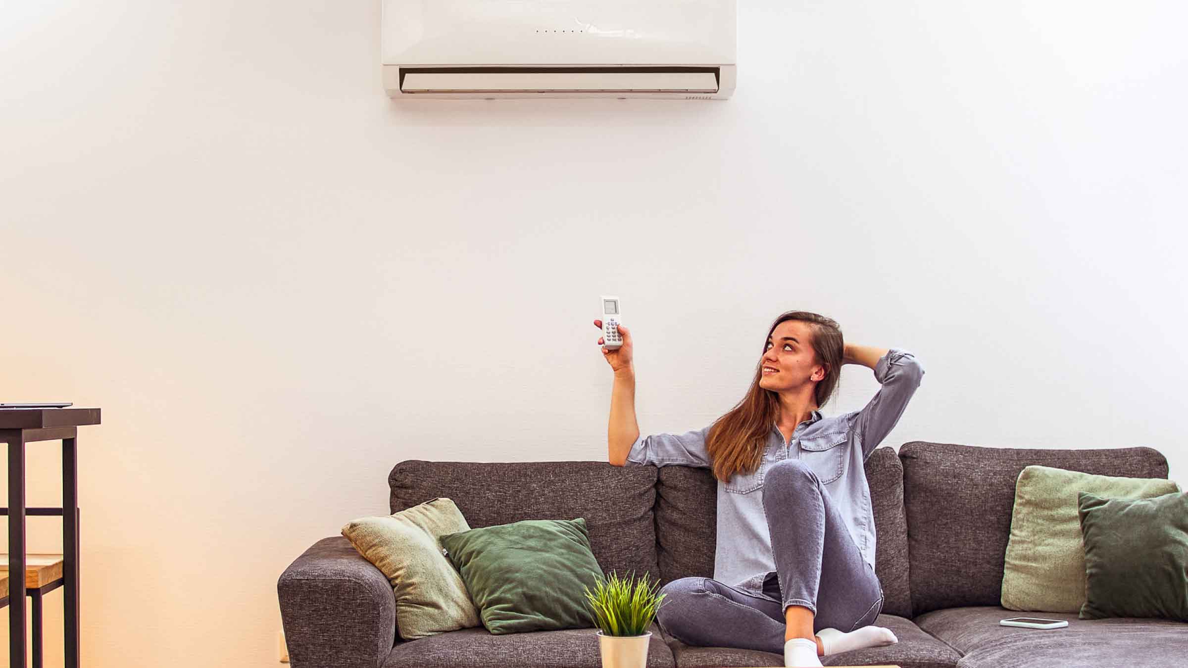 Woman sits on sofa, with her legs up. Looking up towards an air conditioner, with the remote control in her hand.