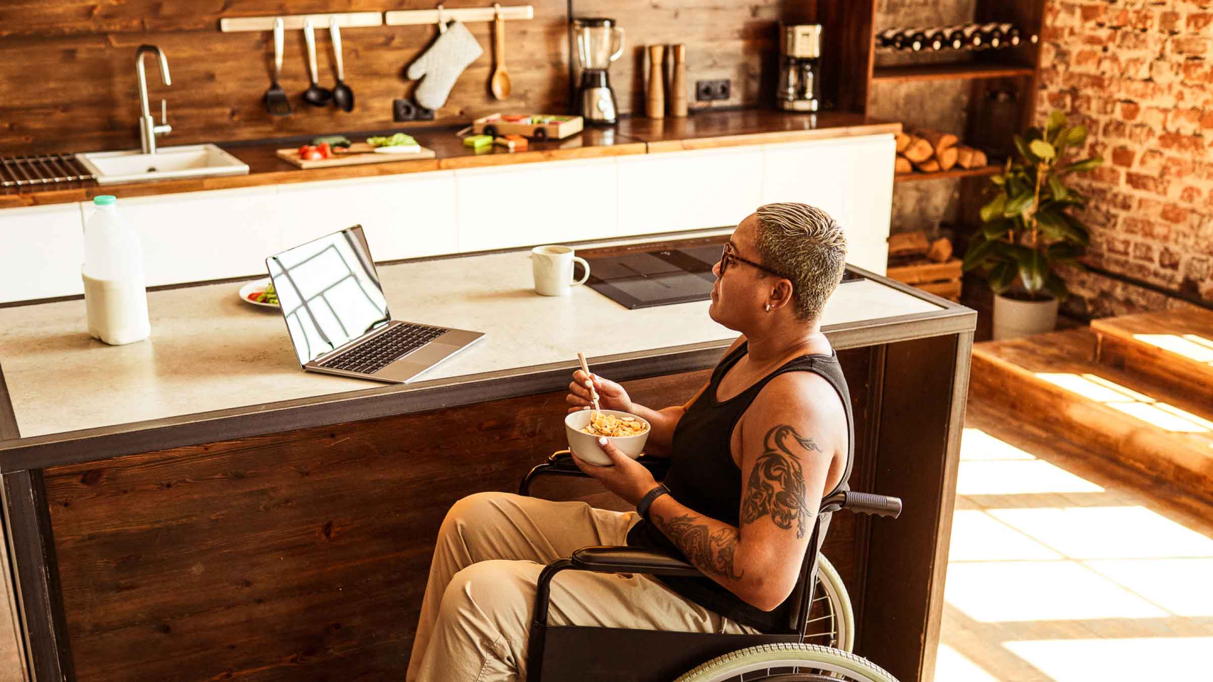 Woman in wheelchair eating cornflakes while watching her laptop perched on kitchen counter. Sun drenched modern kitchen with electric stove top.