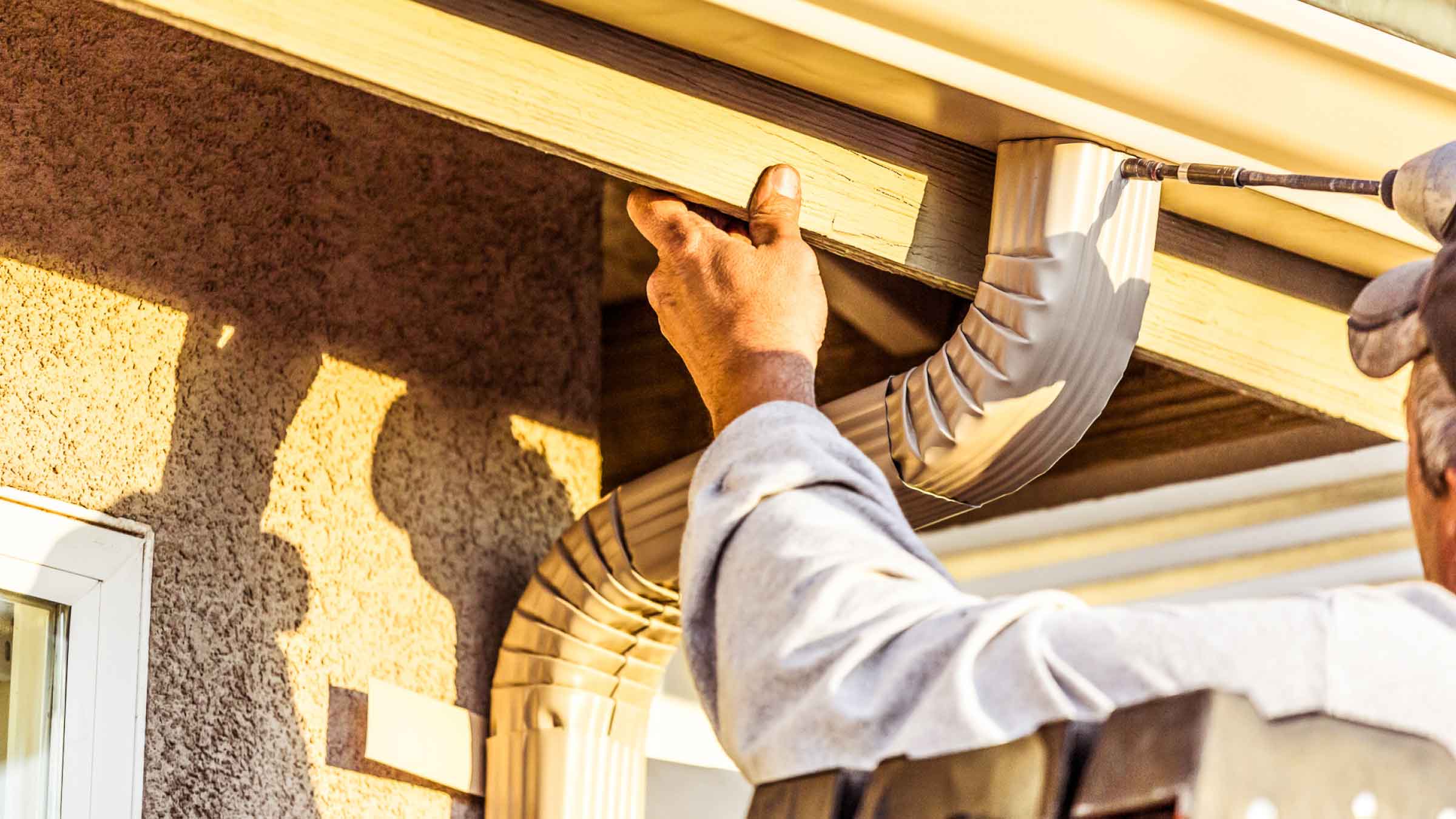 Tradie in a blue shirt, holds onto a timber slat with his left hand while drilling a gutter spout onto the roof with his right hand.