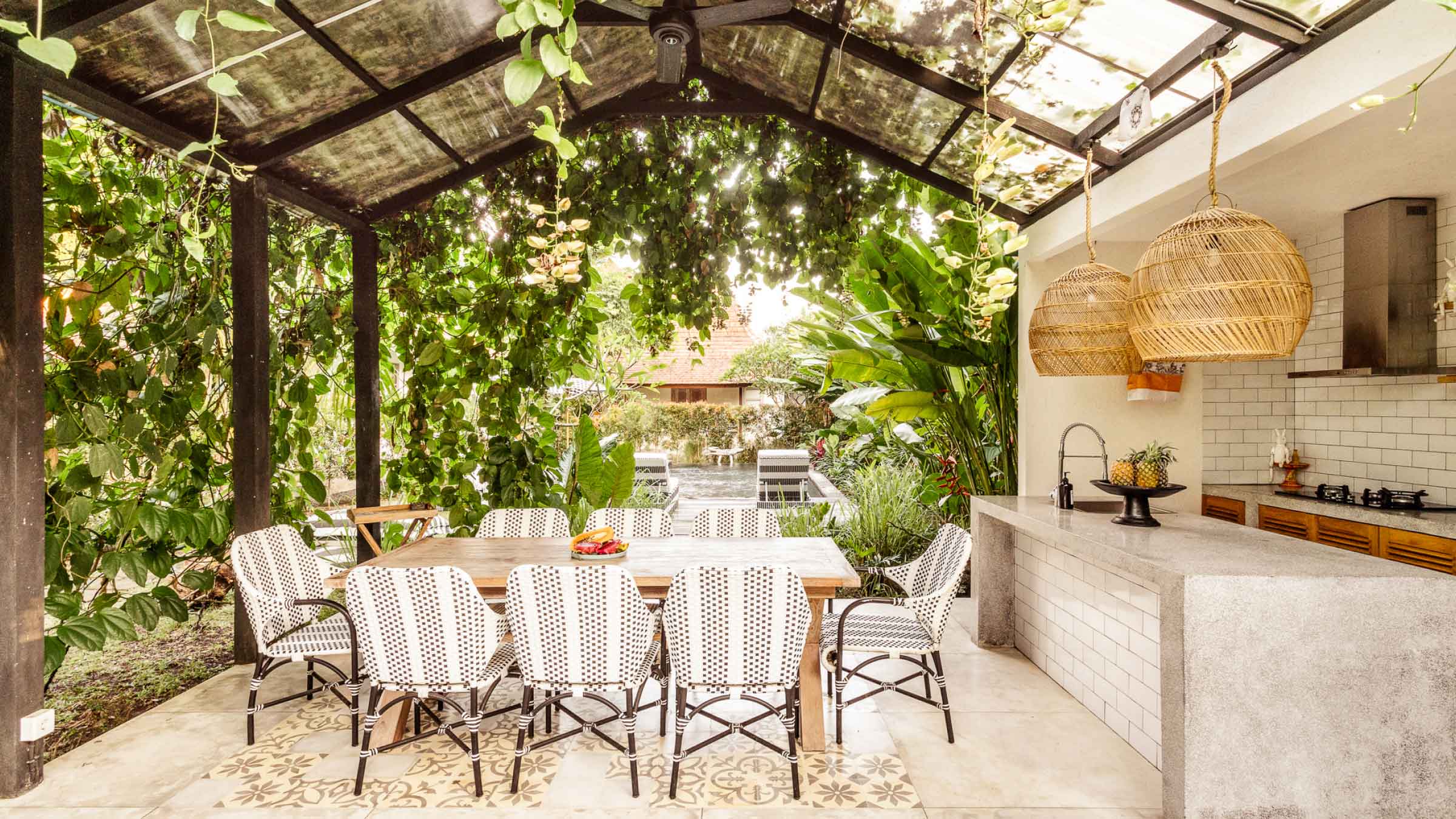 Sunlit outdoor entertainment area, with table and chairs. Under a glass awning with a green leafy canopy.