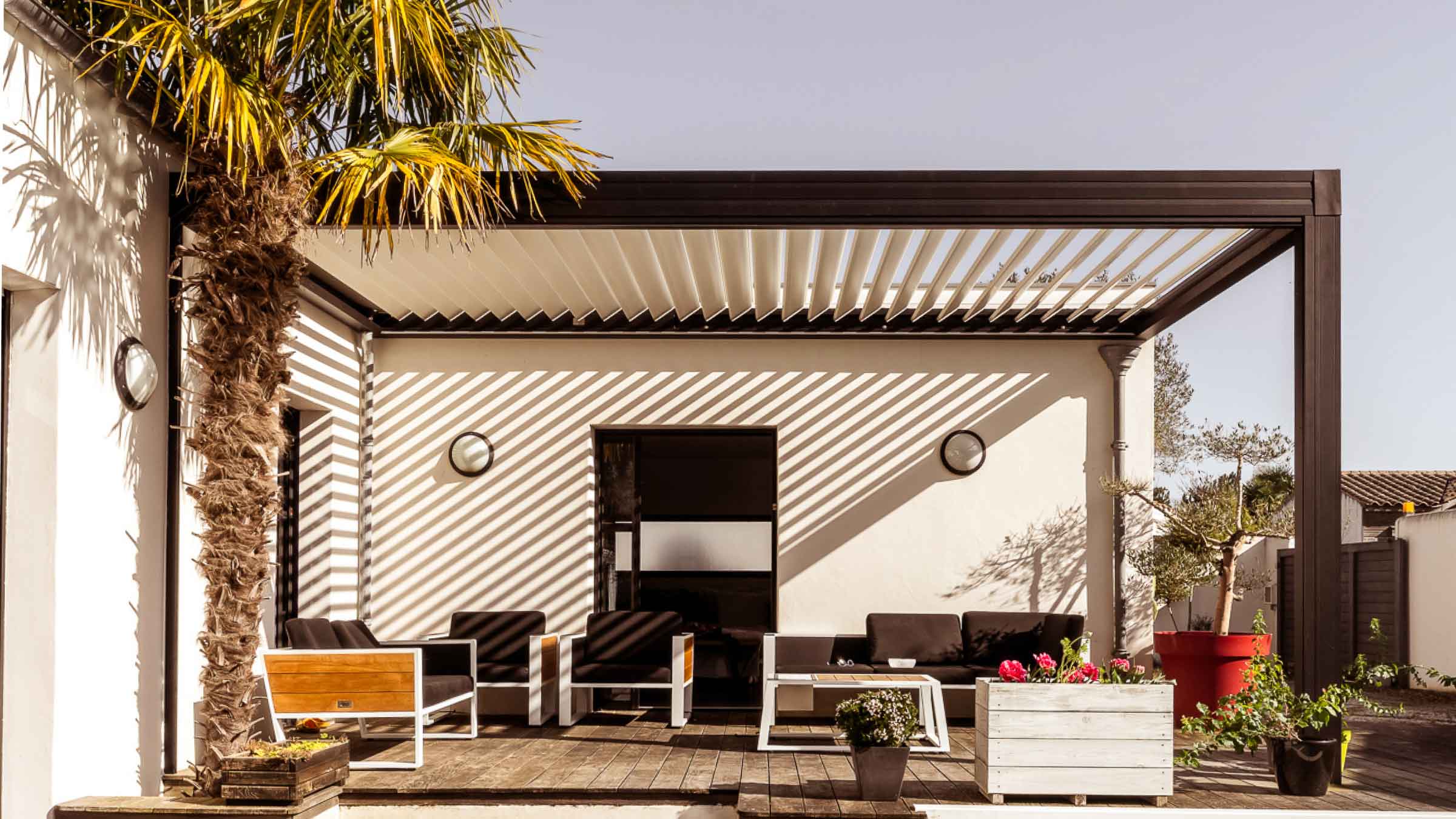 Outdoor entertainment area, with a metal frame surround holding up slotted blinds, which are open allowing sunlight to cast shadows on the white surrounding walls.