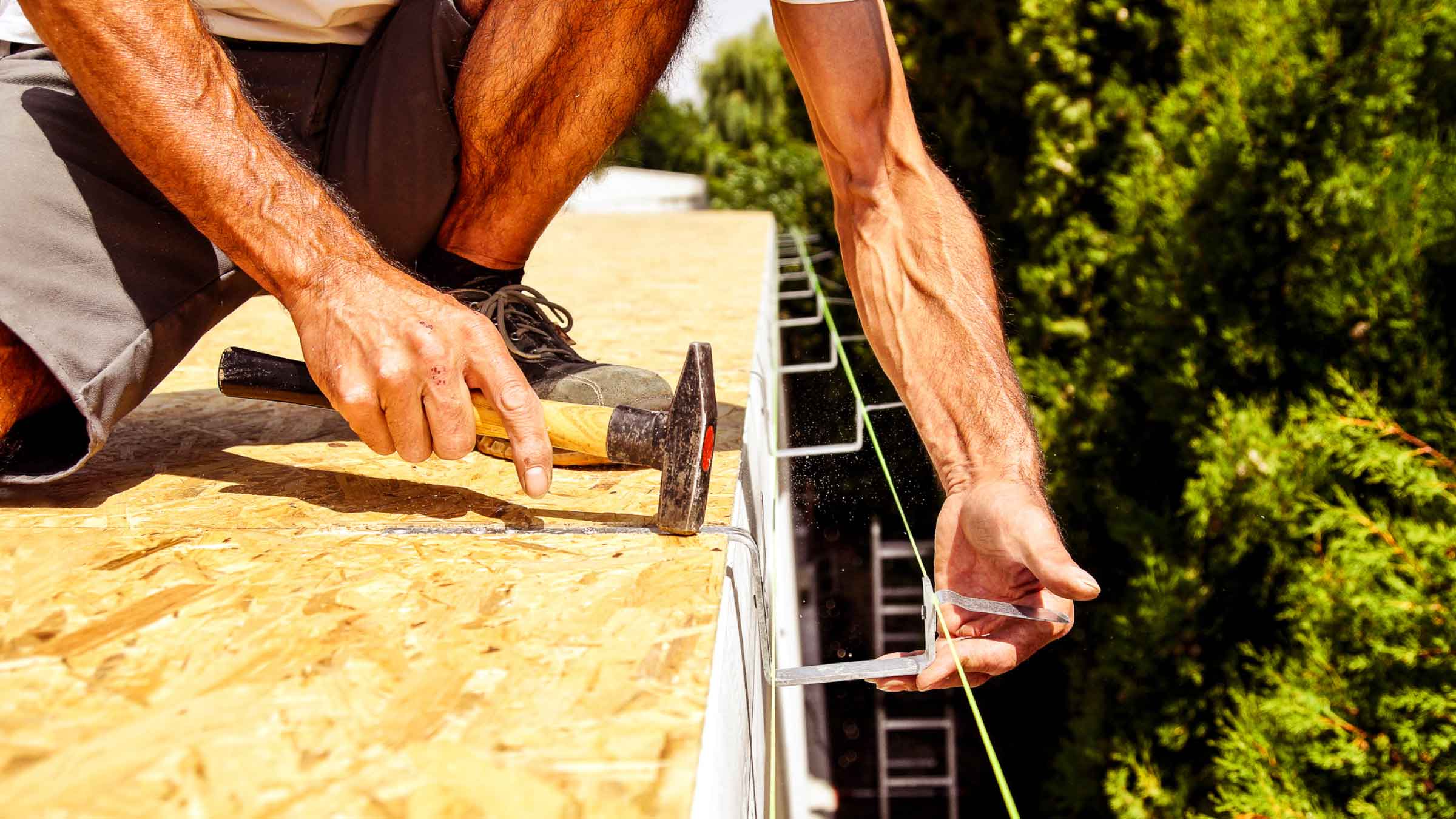 Tradie kneels on a rooftop with a hammer in his right hand as he sets up hanging brackets for a gutter.