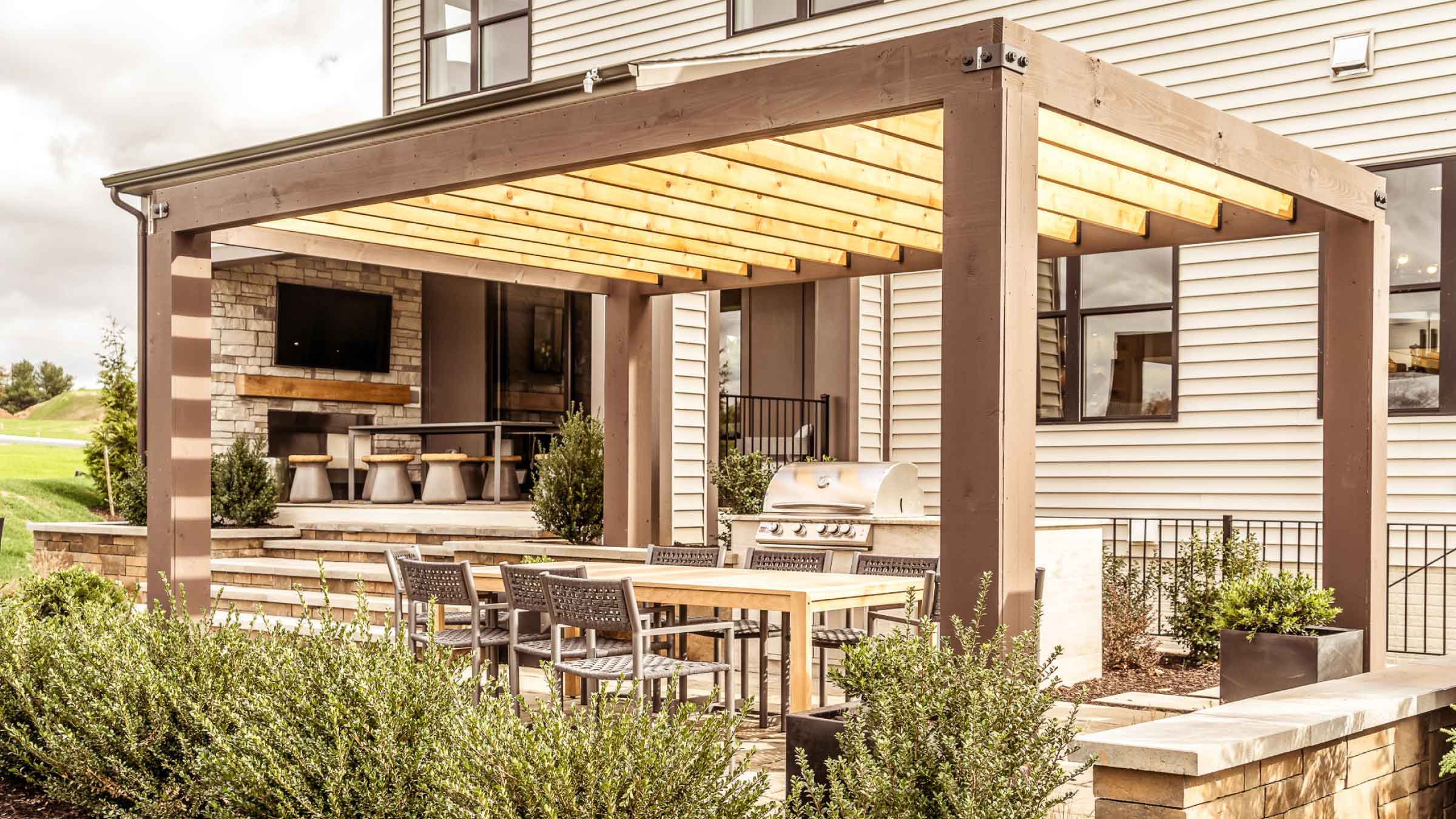 Outdoor entertainment area, made from timber. House in the background, green leafy shrubs dotted around.