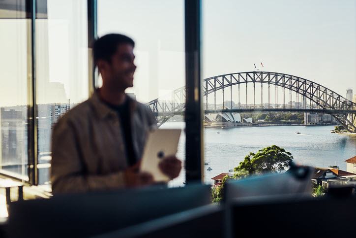 Moneypenny Production Accounting. Photo of person holding tablet blurred in foreground with Sydney harbor in focus outside window in background