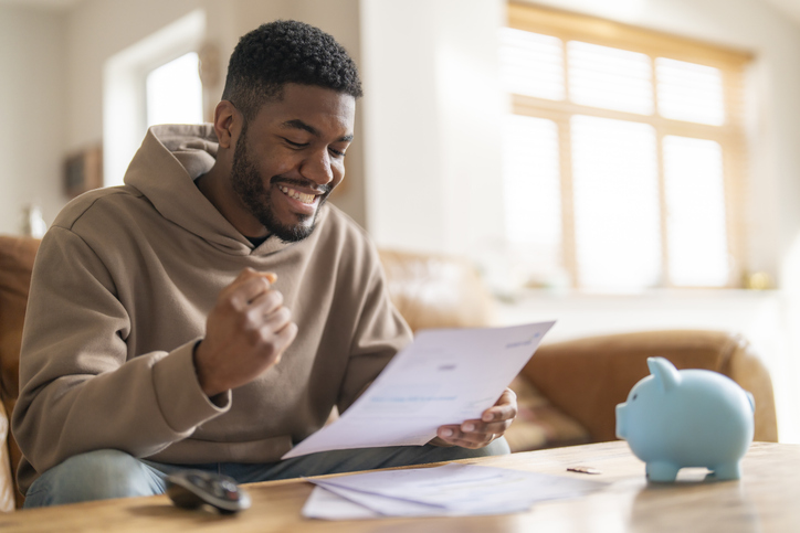 Happy man looking at paperwork in his hand with excited smile. Piggybank on table in front of him.