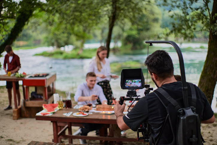 Camera operator filming meal scene at picnic table next to river