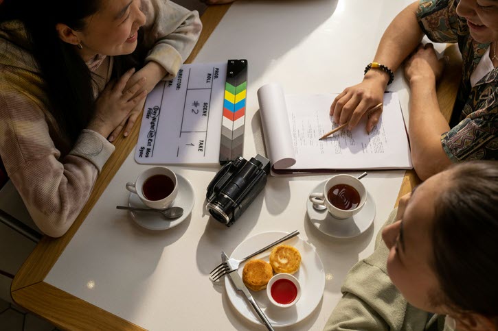 Moneypenny Contracts. Overhead photo of three people at table with production clapboard, script, camera, coffee, pastries on table.