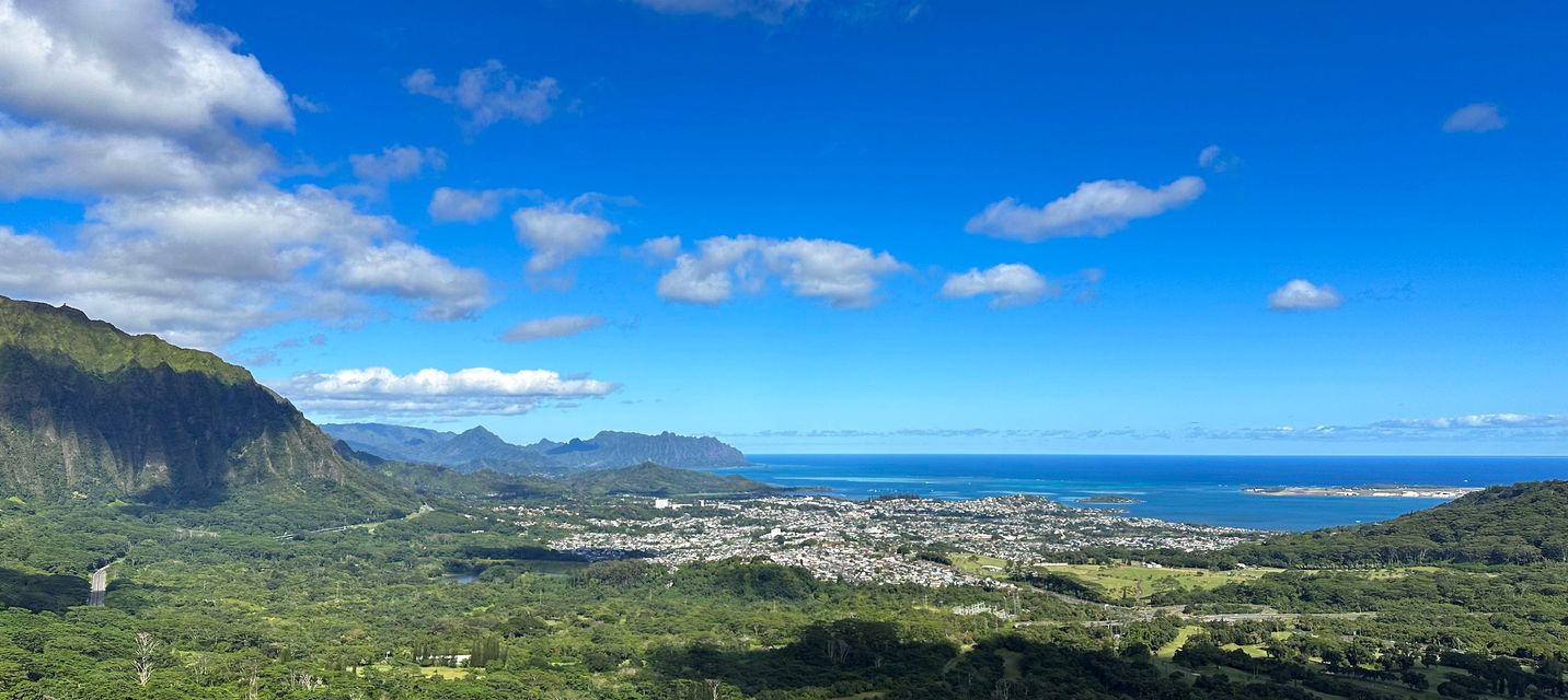 Breathtaking view from the Nuuanu Pali Lookout in Oahu, Hawaii, showcasing lush green valleys, the Koolau Mountains, and the blue ocean in the distance under a bright sky with scattered clouds.