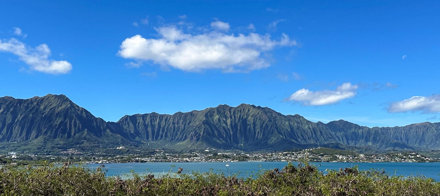 Scenic view of the Koolau Mountain Range in Kaneohe, Hawaii, with lush green peaks, a blue sky dotted with clouds, and calm waters in the foreground.