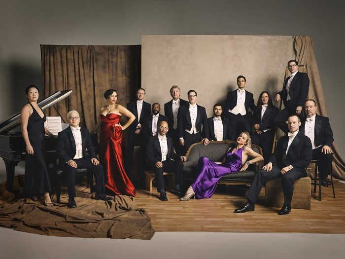 Pink Martini posing in a studio against a brown and tan backdrop. 