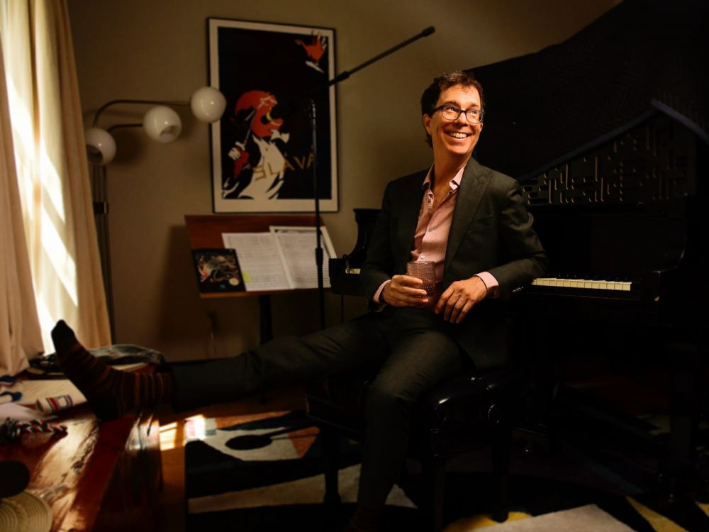 Ben Folds smiling on a piano bench holding a glass in a music room.