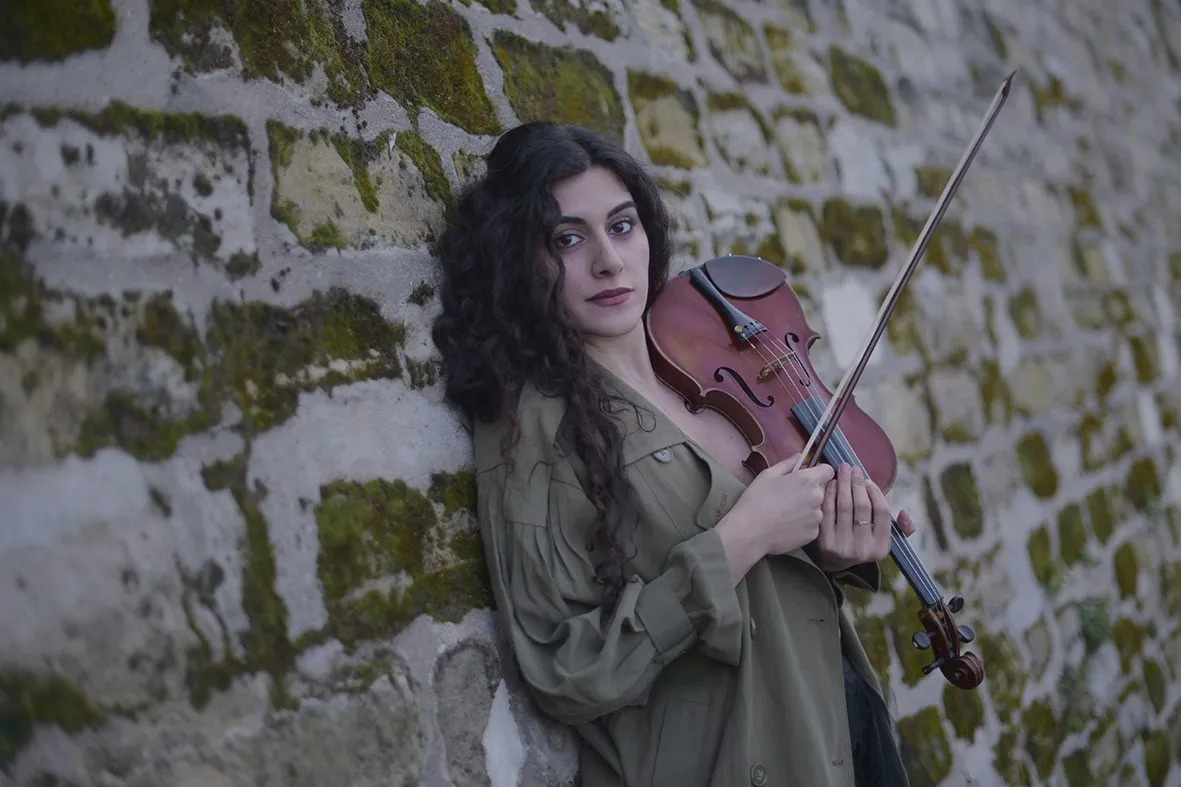 Layle Chaker leaning up against a mossy stone wall holding a violin and bow. 