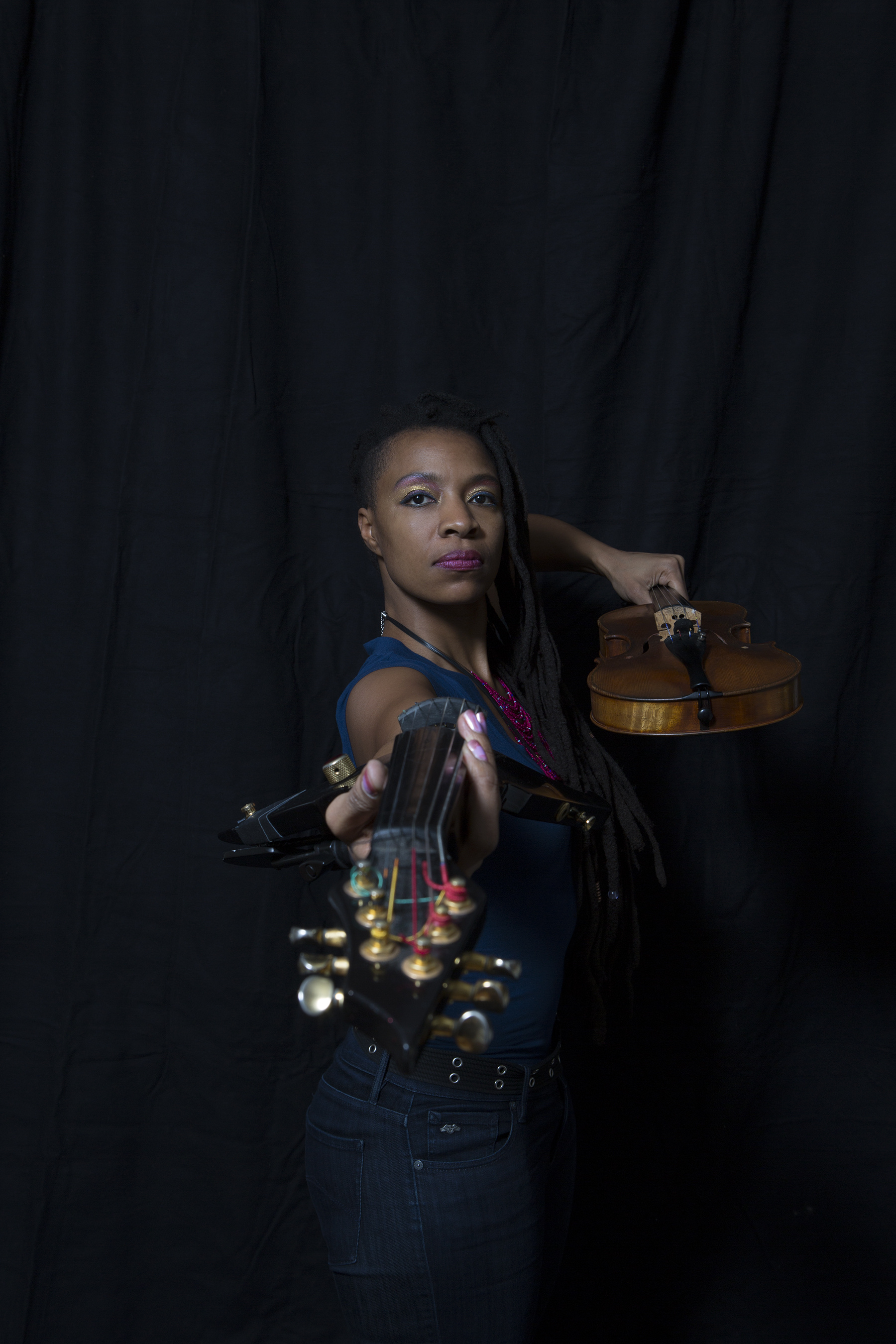 Mazz Swift holding a violin and the neck of a string instrument with a black backdrop. 