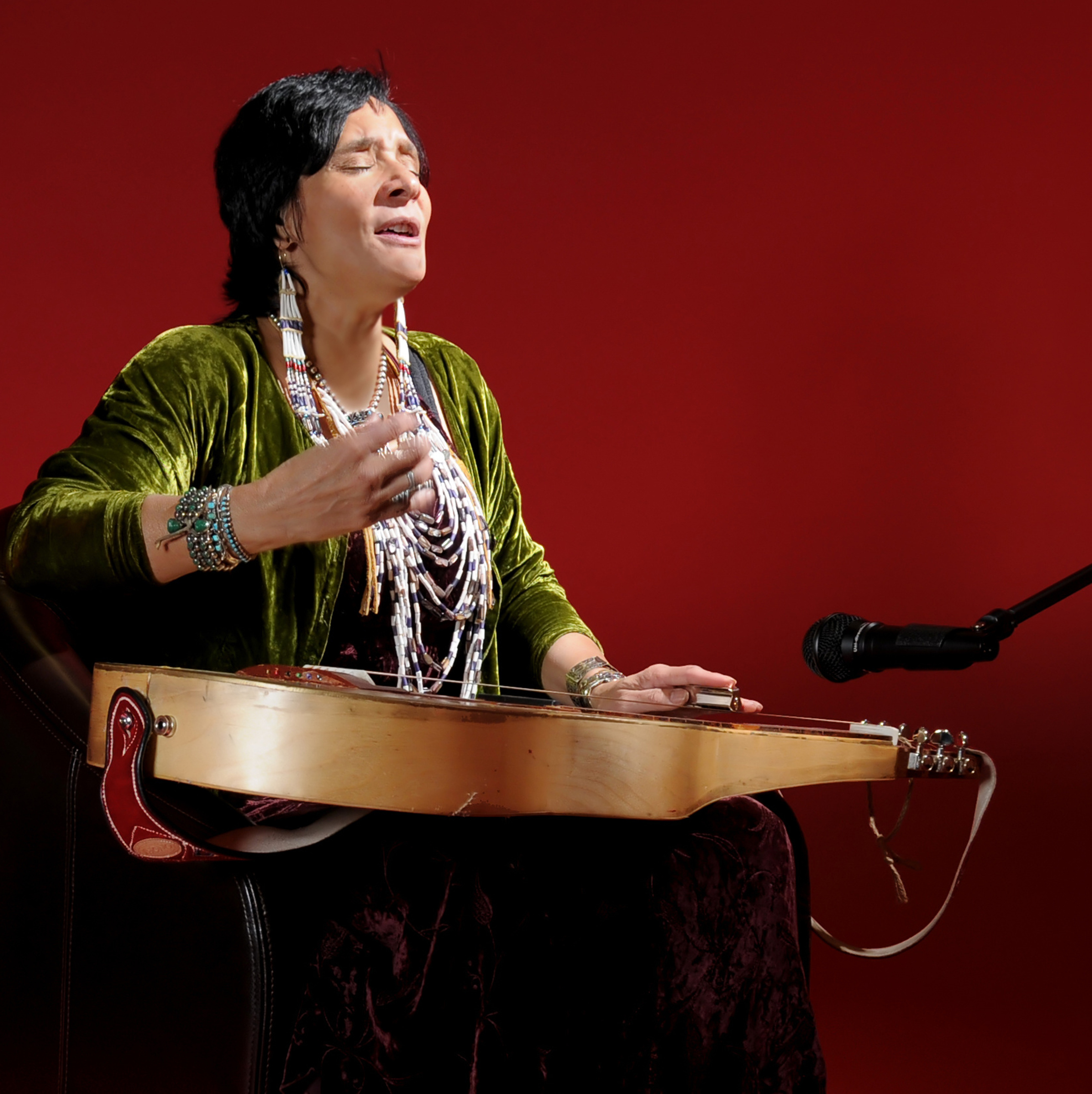 Artist Pura Fé singing and playing a lap-steel slide guitar with a red backdrop. 