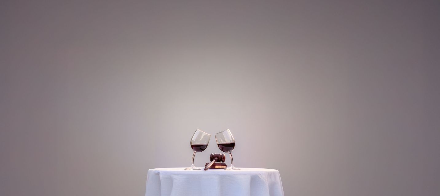 A small round table draped with a white tablecloth, set against a minimalist gradient background. On the table, two glasses of red wine lean toward each other, touching at an angle. Between them, a wooden judge’s gavel rests on a small base, symbolizing a connection between wine and law or justice.