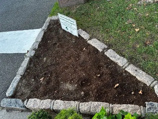 A small, triangular garden bed bordered by stone blocks, freshly prepared with soil but not yet planted. A sign at the back indicates it is part of a local gardening initiative. The surrounding area includes grass, pavement, and a crosswalk.