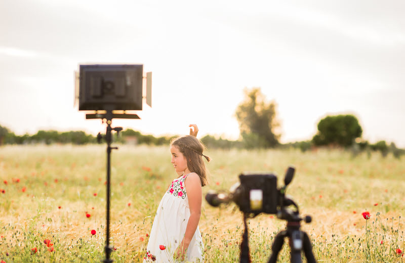 Young actor in a white dress filming in a flower field with professional lighting equipment and a camera on a tripod