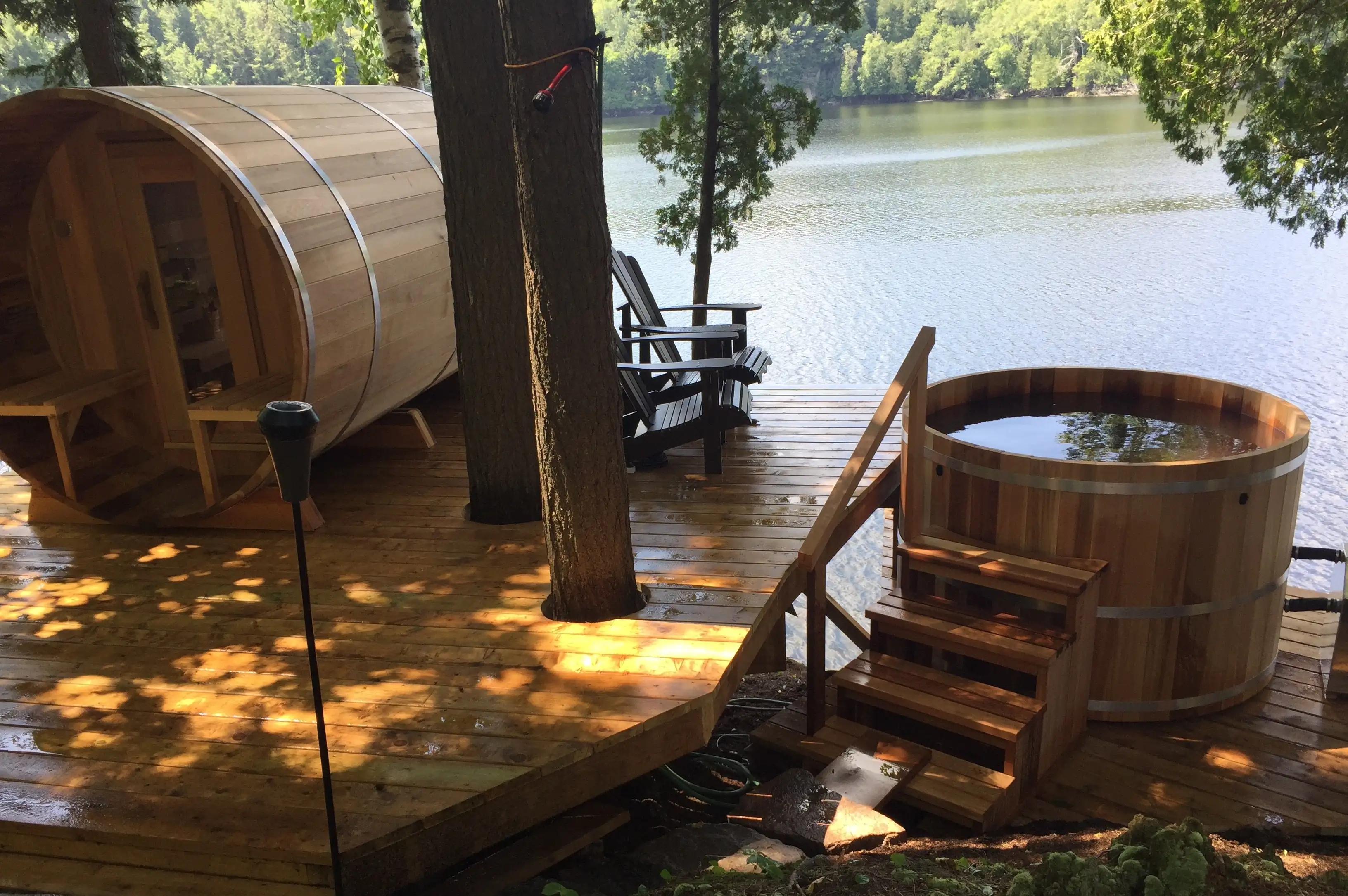 A LeisureCraft cedar barrel sauna and a round cedar cold plunge tub on a lakeside deck, framed by summer forest — the hot and cold sides of the tradition.