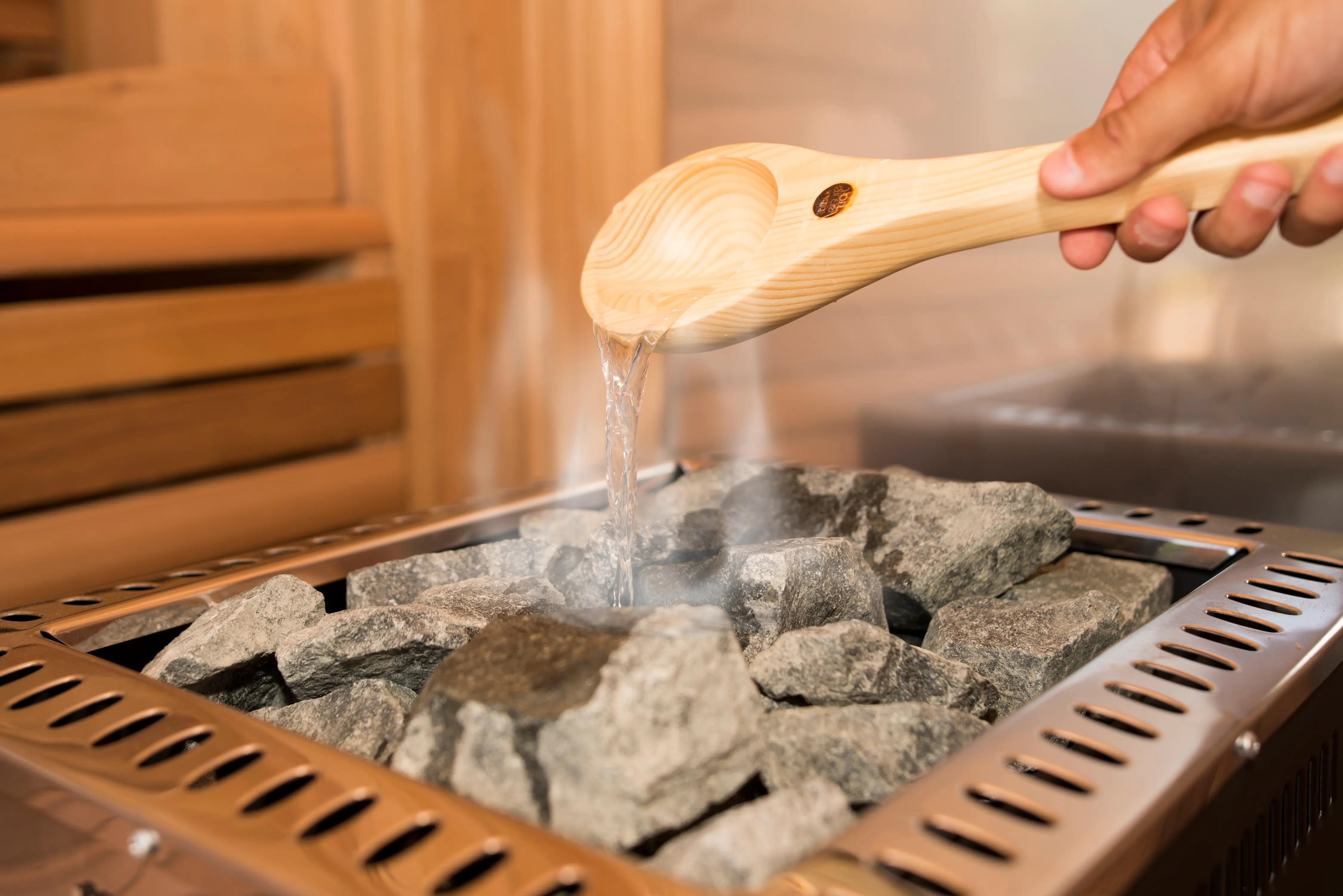 A wooden ladle pouring water over the hot stones of a sauna heater, raising a wisp of steam — the act of making löyly.