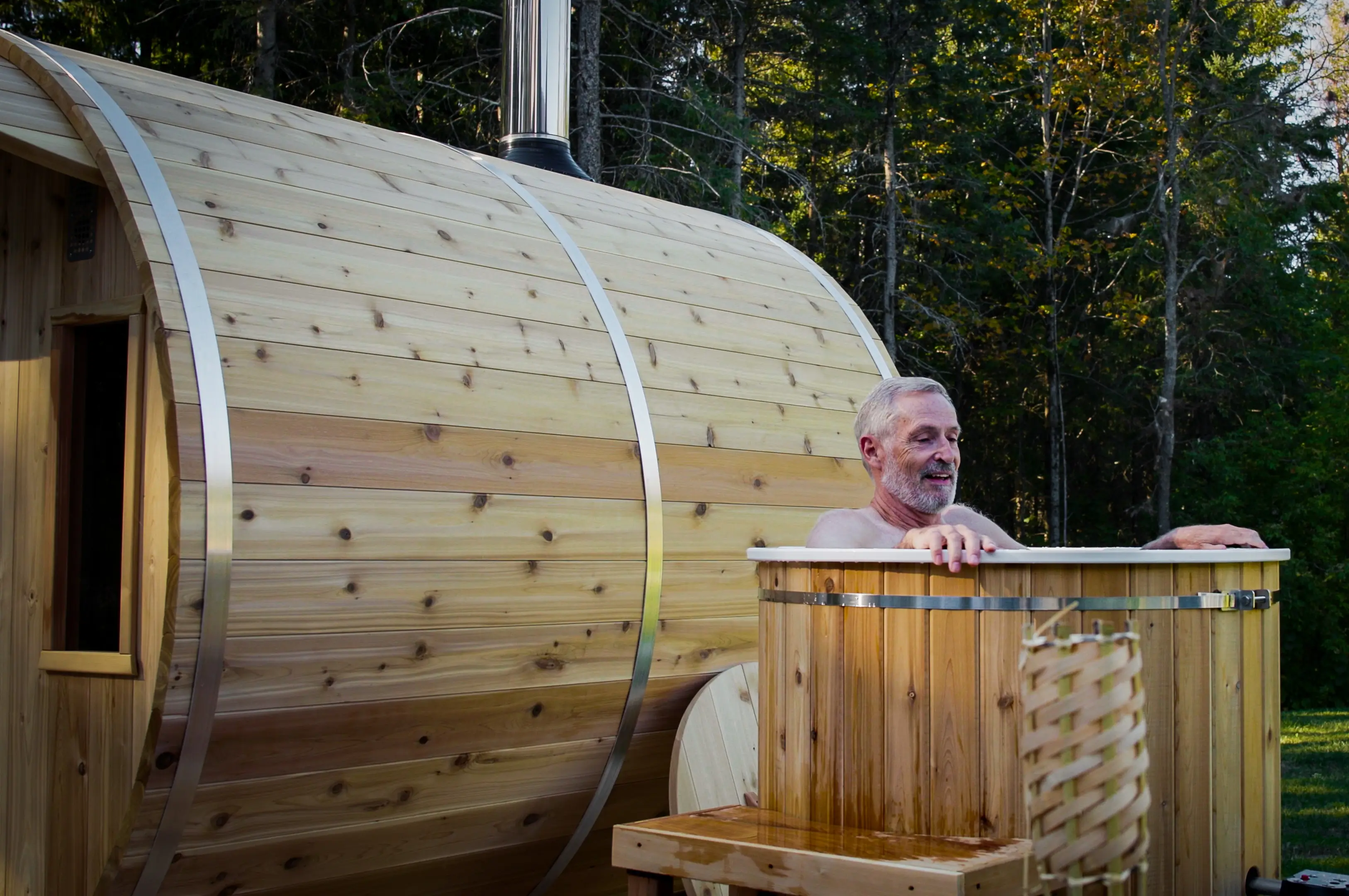 A man relaxing in a cedar cold plunge outdoors, with a barrel sauna beside it