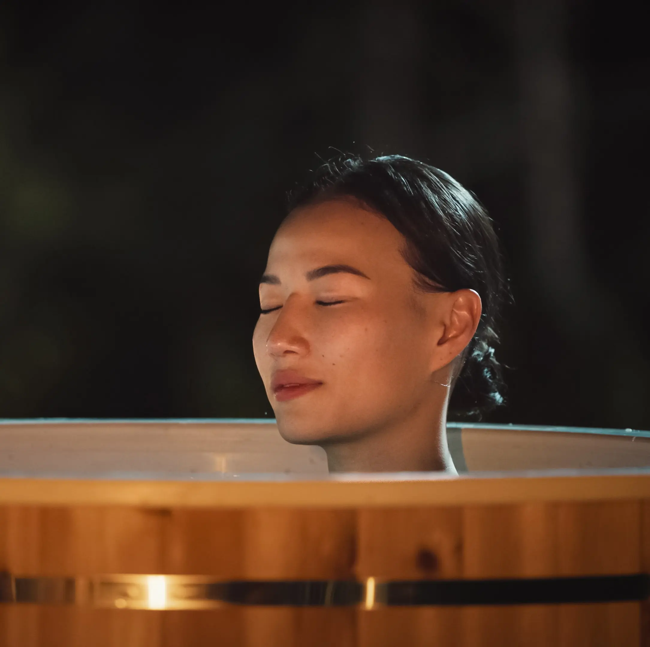 A woman relaxing with her eyes closed in a cedar cold plunge at night