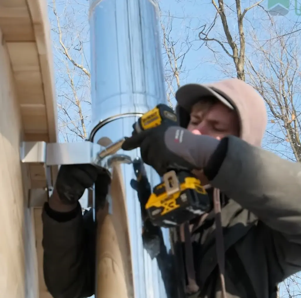 A person adjusting the bracket that secures the stainless steel chimney on an outdoor wood-burning sauna in winter