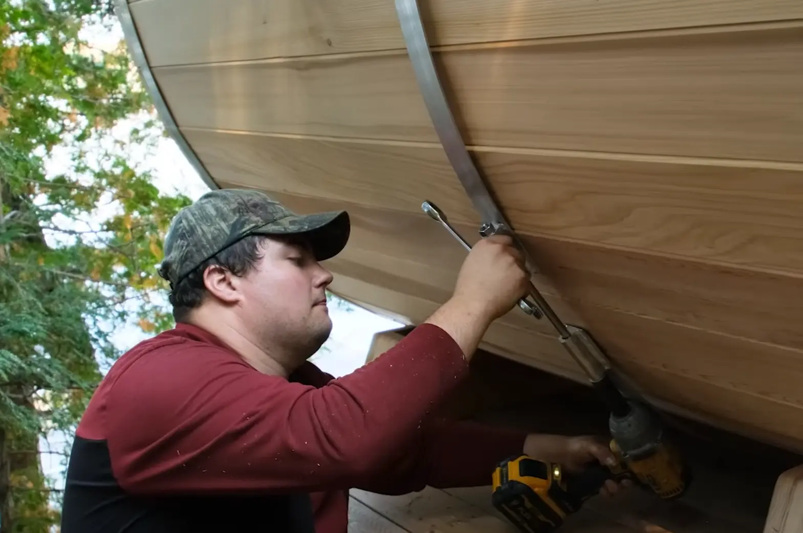 A man tightening the band on a cedar barrel sauna using a cordless drill and a wrench, outdoors