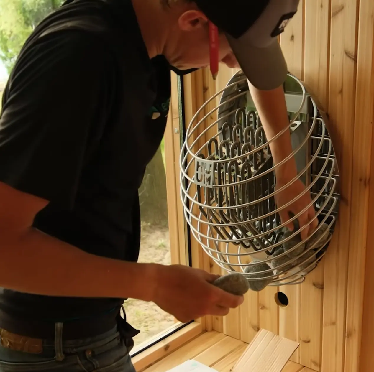 A man placing sauna stones onto a round electric sauna heater inside a cedar sauna