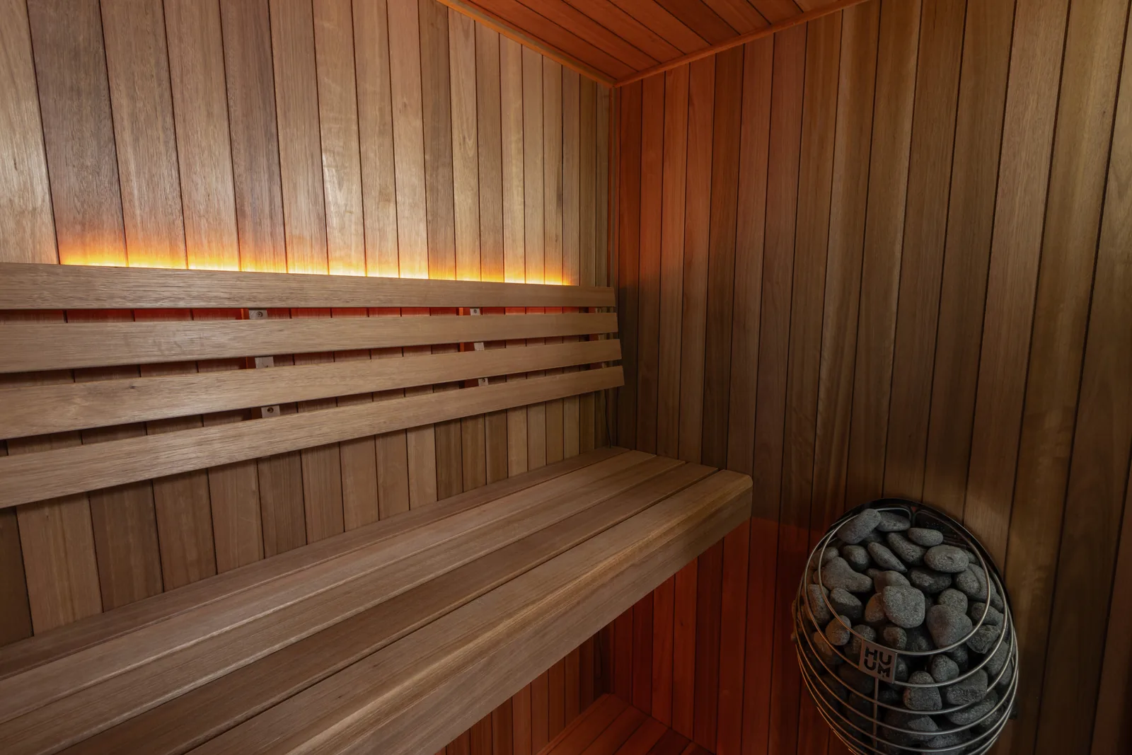 Interior of a LeisureCraft Element indoor sauna in thermally modified Thermo Grandis, with slatted benches, a backrest, an ambient light strip, and a HUUM heater filled with sauna stones.