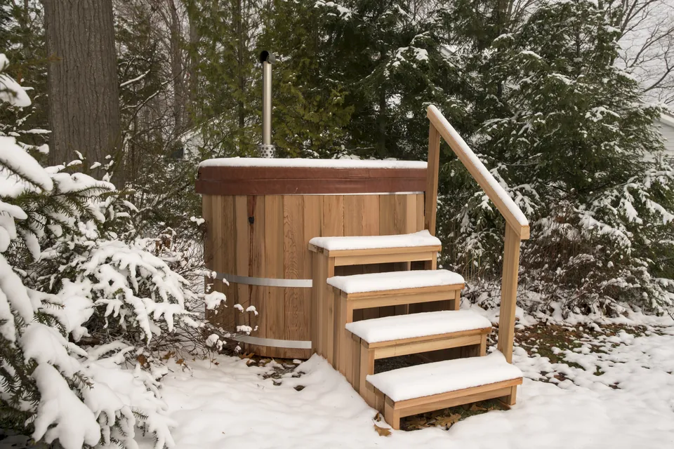A wood-fired hot tub under its insulated winter cover, snow accumulated on top, chimney venting through a cutout