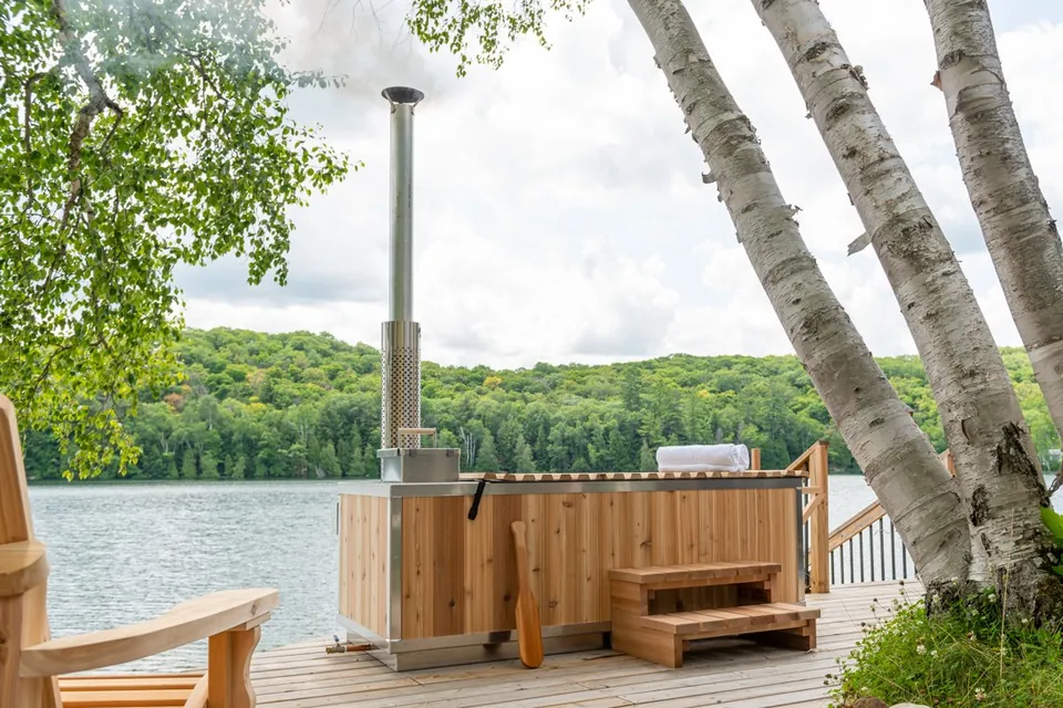 Dundalk Cascade wood-fired hot tub on a lakeside dock among birch trees, chimney venting