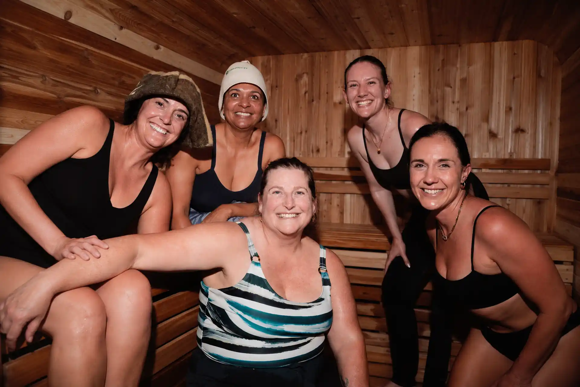 Five women enjoying a Luna Sauna together, laughing and relaxed, some seated and some standing, a couple wearing sauna hats