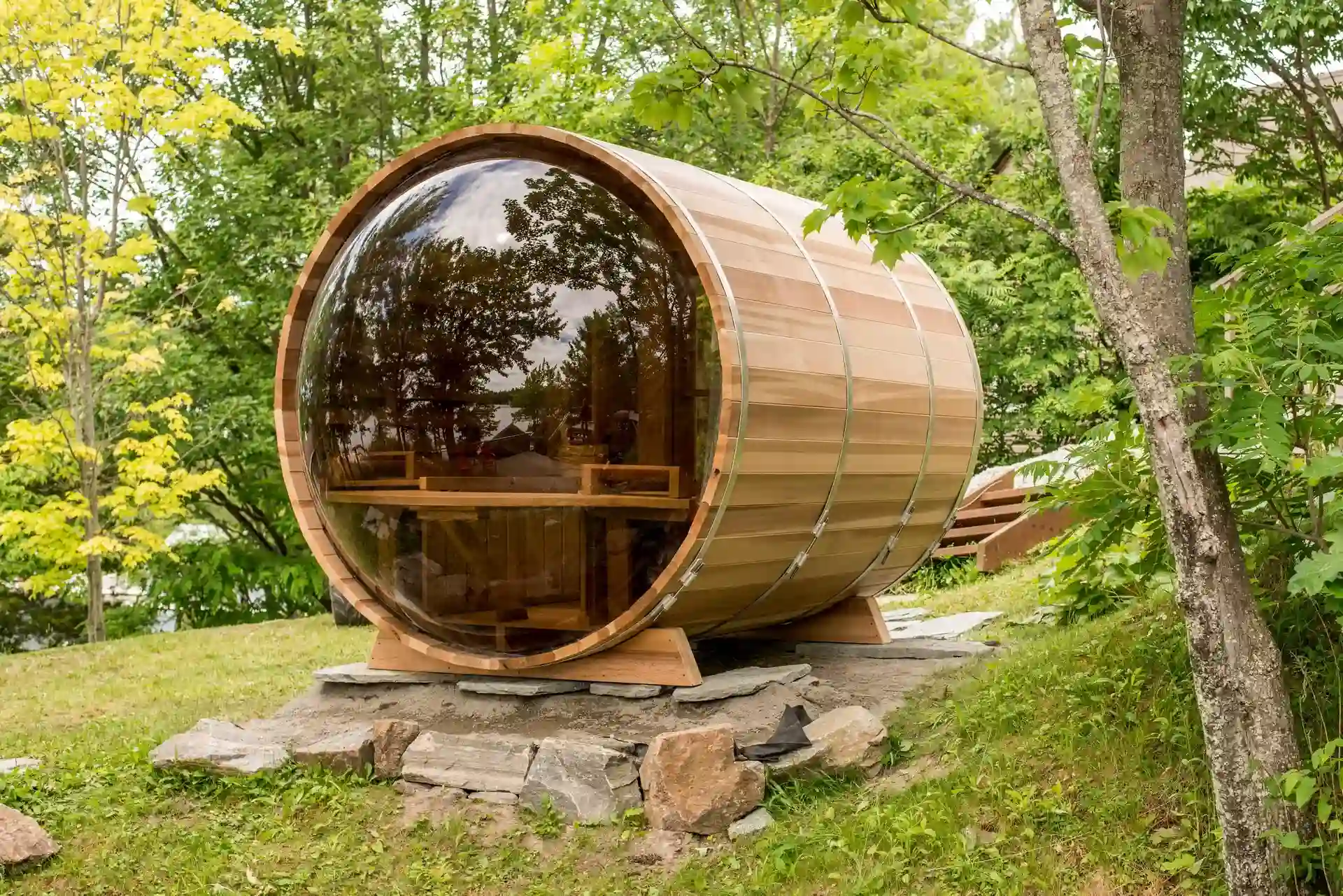 Panoramic barrel sauna in western red cedar with stainless steel bands, set on a stone foundation in a wooded Canadian backyard