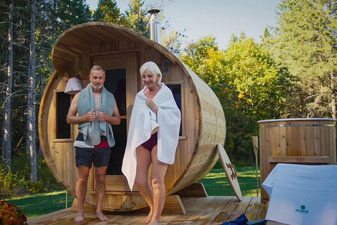 An older couple stepping out of a LeisureCraft cedar barrel sauna onto a wooden deck, with a cedar cold plunge tub alongside and autumn woods behind
