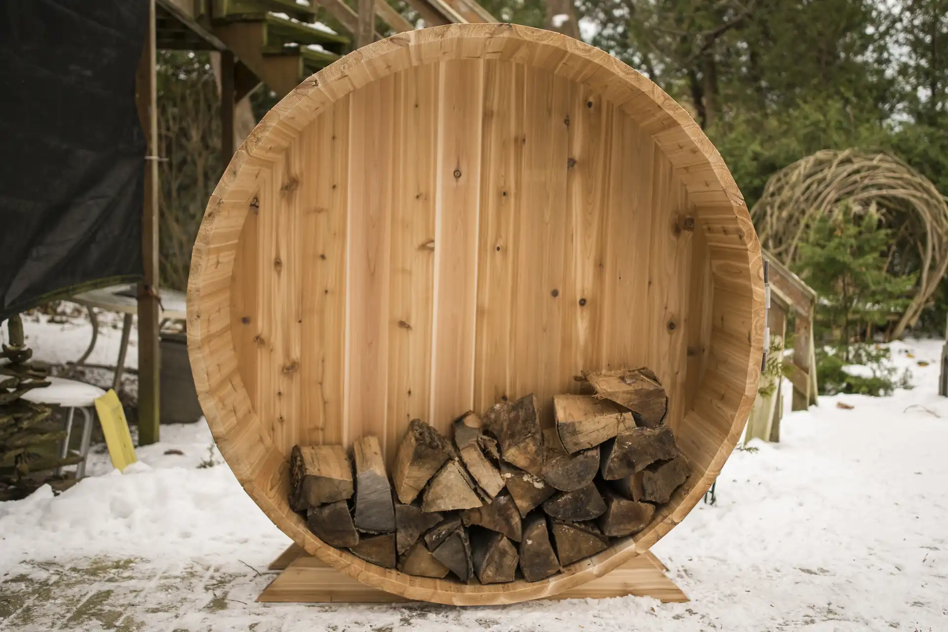5 foot knotty cedar firewood storage barrel filled with split hardwood on a snowy backyard patio