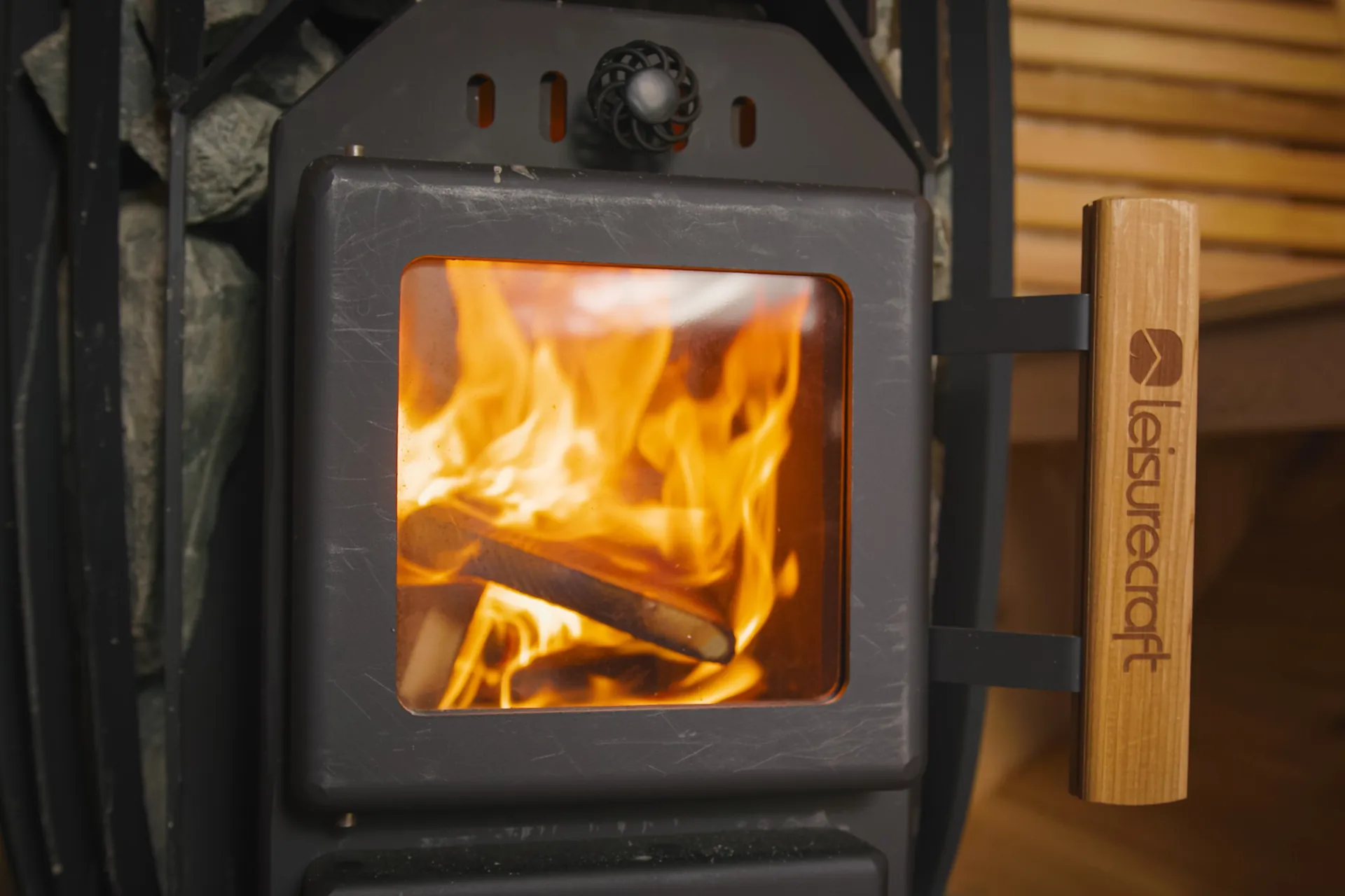 Fire burning behind the glass door of a TimberGlow14 wood-burning sauna heater, with sauna stones and a LeisureCraft cedar door handle