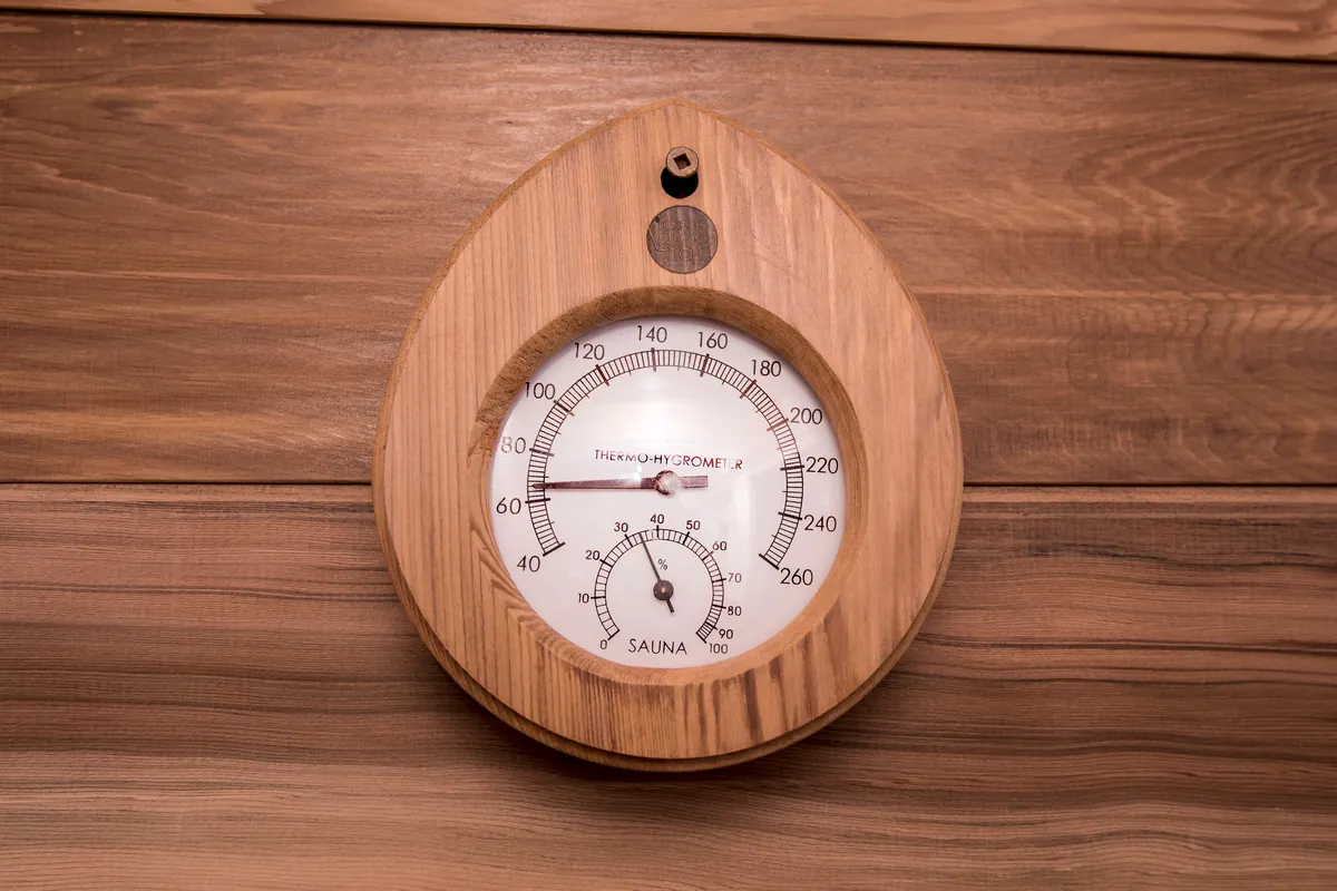 Teardrop-shaped wooden sauna thermo-hygrometer on a wood-paneled wall, with a Fahrenheit temperature dial, a smaller humidity dial, and a Dundalk LeisureCraft badge