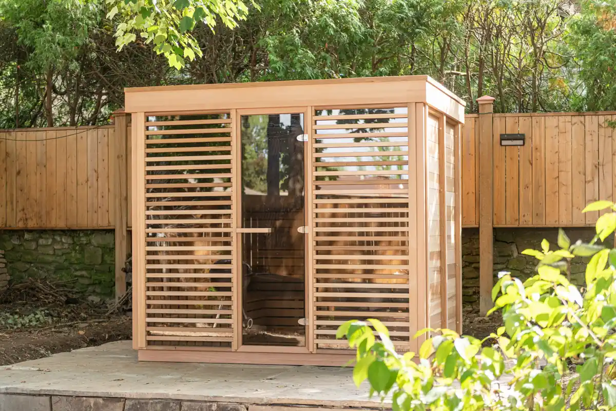 Cedar cube sauna with a glass door between two semi-privacy panels of horizontal cedar slats and a solid cedar side wall, set on a stone patio by a wooden fence
