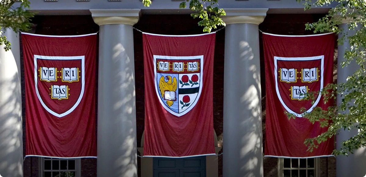 Front steps of a Harvard University building with large columns and red banners displaying the Harvard crest; groups of people are gathered on the stone steps beneath trees.