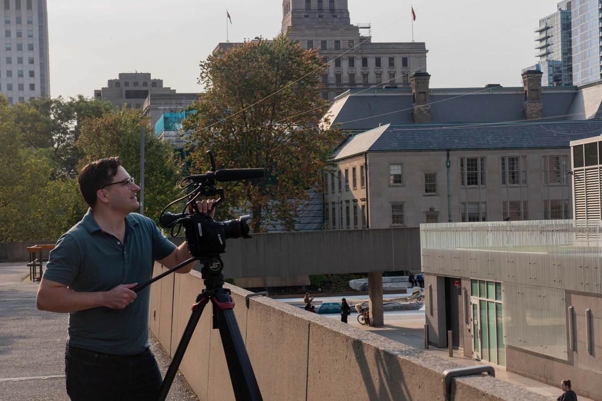 A man in a blue shirt stands on a rooftop, filming with a camera.
